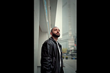 A man with a closely shaved head looks upward while standing on a city street, tall glass buildings rising behind him.