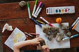 An aerial view of a markers and paints spread out on a wooden table. You can see a child’s hands painting on rocks and paper.