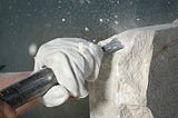 A white gloved left hand is holding a chisel to a piece of limestone. At the point where they meet, there is a cloud of stone chips and dust, as though the chisel has just been hit by a hammer.