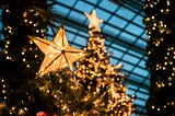 A close-up of a Christmas tree with a star covered in white lights in the foreground with a glass ceiling, a second Christmas tree, and two pillars wrapped in garland and lights in the background.