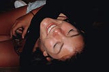 A close-up of a young woman on the floor, eyes closed and smiling skyward. She’s holding sunglasses in one hand and also looks to be intoxicated.