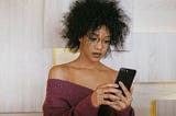 A beautiful African American woman with curly black hair wears round glasses in brown attire and reads on her iPhone.