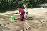 Photo of two of the author’s young children playing with a hose and bucket of water on a driveway.