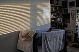 The image shows a baby travel bed in a dark room with light reflections from the window blinds on a white closet.