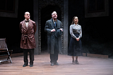 Three actors stand onstage and prepare to take a bow; the actor on the left wears a patterned smoking jacket, the one in the middle a gray suit, and the one on the right a blue skirt and floral blouse.