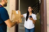 A smiling woman receives a food delivery at her doorstep from a courier holding a paper bag, representing the convenience and risks of home-delivered meals.