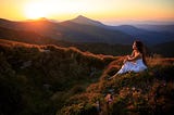 A lady in white dress sits on the earth staring at the setting sun