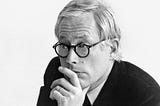 A photo of designer Dieter Rams. He is wearing horn rimmed glasses and has his hand on his chin in a thoughtful pose.