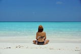 Woman mindfully sitting cross-legged on a beach, looking out at ocean.