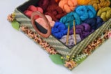 A rectangular box filled with many colours of yarn and two gold crochet hooks. The box has a round wooden handle and there are leaves and dried flowers all around the bottom.