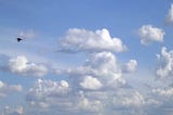 Um urubu no céu azul infestado de nuvens brancas e cinzas, ele está um pouco depois da metade da imagem do lado esquerdo da imagem batendo as asas, o clique pegou elas inclinadas para baixo.