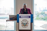 Cheryl Andrew-Maltais of the Aquinnah Wampanoag Tribe of Gay Head, a woman with long gray hair in a side braid, speaks at a podium with a banner that says, “Welcome. Massachusetts Tribal and Indigenous Health Summit 2023 Department of Public Health”