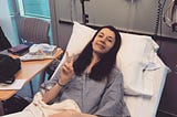Summer, a woman in her early twenties, smiles from a hospital bed. She wears a gown and her hand makes a “peace” sign.