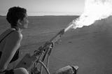 A short-haired woman in a tank top straddles a fire cannon, blasting a puff of flames into the sky in front of a desert horizon.