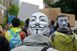 A photo taken from the back of a crowd picketing for some unknown cause. The photo is taken directly from behind someone wearing a black-and-white Guy Fawkes mask on the back of their head, which is facing the camera. There is another person with the same mask on the back of their heads to the front and right of the first person. Several other people can be seen in front, and building and trees further ahead to either side.