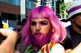 A man with a purple beard and shoulder-length hair with rainbow-colored earrings and a rainbow necklace looks at the camera among a crowd of people