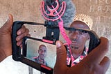 Hands of a citizen reporter holding a mobile camera surrounded by a stabilising rig and a microphone attachment. In the camera viewfinder is an image of a Sierra Leonean man waiting to be interviewed.