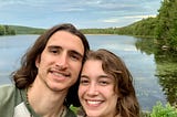 My partner and I smiling in front of a lake surrounded by green, bushy trees.