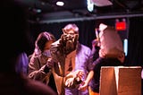 In a dark room, a participant examines a cardboard mask with a triangular nose and textured tufts of hair standing up. In the background, people try other masks.