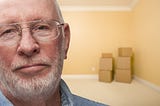 older white man looking sad with boxes in corner of empty room