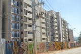 Prédio residencial de vários andares com fachada cinza claro, localizado atrás de uma cerca de metal colorida com grafites. Em primeiro plano, há um poste de eletricidade com fios e transformadores suspensos que cruzam a imagem. À direita, uma rua asfaltada acompanha a lateral do condomínio, que se estende ao fundo sob um céu azul e limpo.