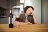 A woman with head in her hand looking stressed out with a glass of wine and a bottle next to it.