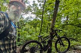 The author, a bearded man wearing a red helmet, glasses, a plaid shirt, and a hydration pack, takes a selfie in a dense, green forest. He appears slightly breathless or surprised. Beside him is a black mountain bike resting against a tree. The trail is rocky and surrounded by lush foliage, indicating a rugged and remote woodland setting.