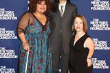 Three women standing against a blue and white backdrop reading “NY Women’s Foundation. One is an African and Indigenous ancestry with curly shoulder length hair and a dress that has sheer black sleeves and a blue flowered pattern. Next to her is an Korean American woman in a grey suit with a grey tie and a white shirt. Next to her is a white, achondroplastic dwarf woman with red hair and freckles wearing a Black sleeveless jumpsuit.