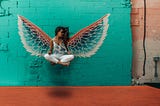 A woman sitting cross-legged, apparently hovering in mid-air but presumably on a hidden, suspended platform. She is sitting against a brick wall with angel wings painted on, and positioned so that the wings appear to spring from her back.