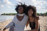 Two beautiful black women embrace each other while smiling on the beach. Their afros blow in the wind as the sun kissed their skin.