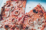 A pizza missing a slice in a box. The pizza is topped with seafood including calamari rings and tiny octopus.