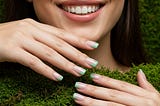 A young woman in her 20s with simple Divine sage green sculpted tips for March 2025, featuring subtle 3D edges molded with clear sculpting gel, hands against a spring moss backdrop — vivid, photorealistic.