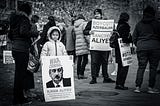 A child holding a sign with “War Criminal: Ilham Aliyev” and a man holding a sign with “Boycott Azerbaijan / Sanction Aliyev”