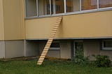 A cat ladder leans against a balcony wall of a Soviet Era style apartment building in Lithuania. The ladder is a board about 4 meters in length with about 15 small slats attached horizontally to act as stairs or steps on the ladder.