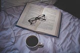 A book set on top a bed with a pair of folded reading glasses on the book. Nearby is a laptop and a cup of coffee.