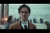 image of the character Syril Karn from the TV show Andor. He is standing in a plaza, looking bewildered, as a fight between fascists and rebels erupts all around him