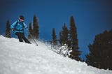Skier in blue ski jacket, black helmet and ski pants, and multicolored ski goggles skiing down a slope with fresh snow against green evergreens and a dark blue sky.