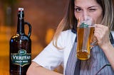 At left is a bottle of Veritas Craft Beer. At right, an attractive female beer hall patron named Polly winks as she drinks from a tall mug, leaning her elbow on the table.