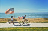 Woman holding American flag high while chasing bike ridden by a man wearing the American flag over his shoulders.