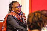 A Kenyan woman with braids and glasses speaks into a microphone
