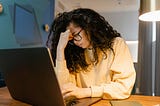 Young woman with dark curly hair holding her head in one hand while typing on her laptop keyboard with the other