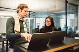 Man and woman stand in front of two laptops on a high table and discuss what is on the screen