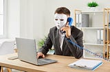 A man wearing a mask sitting at a laptop while he talks on a phone