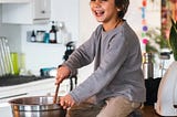 A smiling child sits on a kitchen counter while mixing chocolate batter in a large metal bowl, enjoying a baking moment.