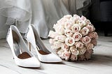 A pair of white wedding shoes on a hardwood floor next to a light pink rose bouquet.