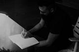 Black and white photo of a man writing in a notebook. The image is dark and faded to create a dramatic effect.