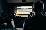 View from inside a conference audience. An out-of-focus attendee sits in the foreground, watching a distant leader on stage present a slide reading “OUR NEW FOCUS: PROFITABILITY & EFFICIENCY.” The auditorium is dim; the screen glows against silhouetted heads.