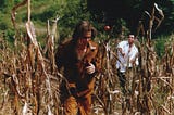 A man dressed like Tom Sawyer runs through a field laughing, as a tomato whizzes through the air, hurled by another man, seen in the distance, wearing a white button up shirt.