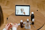Desktop with medicine bottle and a laptop computer displaying a bearded therapist looking at a seated individual.