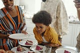 young brown skin boy in center of photo with mustard colored shirt starring at a plate with strawberry cake. a black lady on his right with a blue and orange dress on holding a plate. a man with a white shirt is behind boy, face out of frame